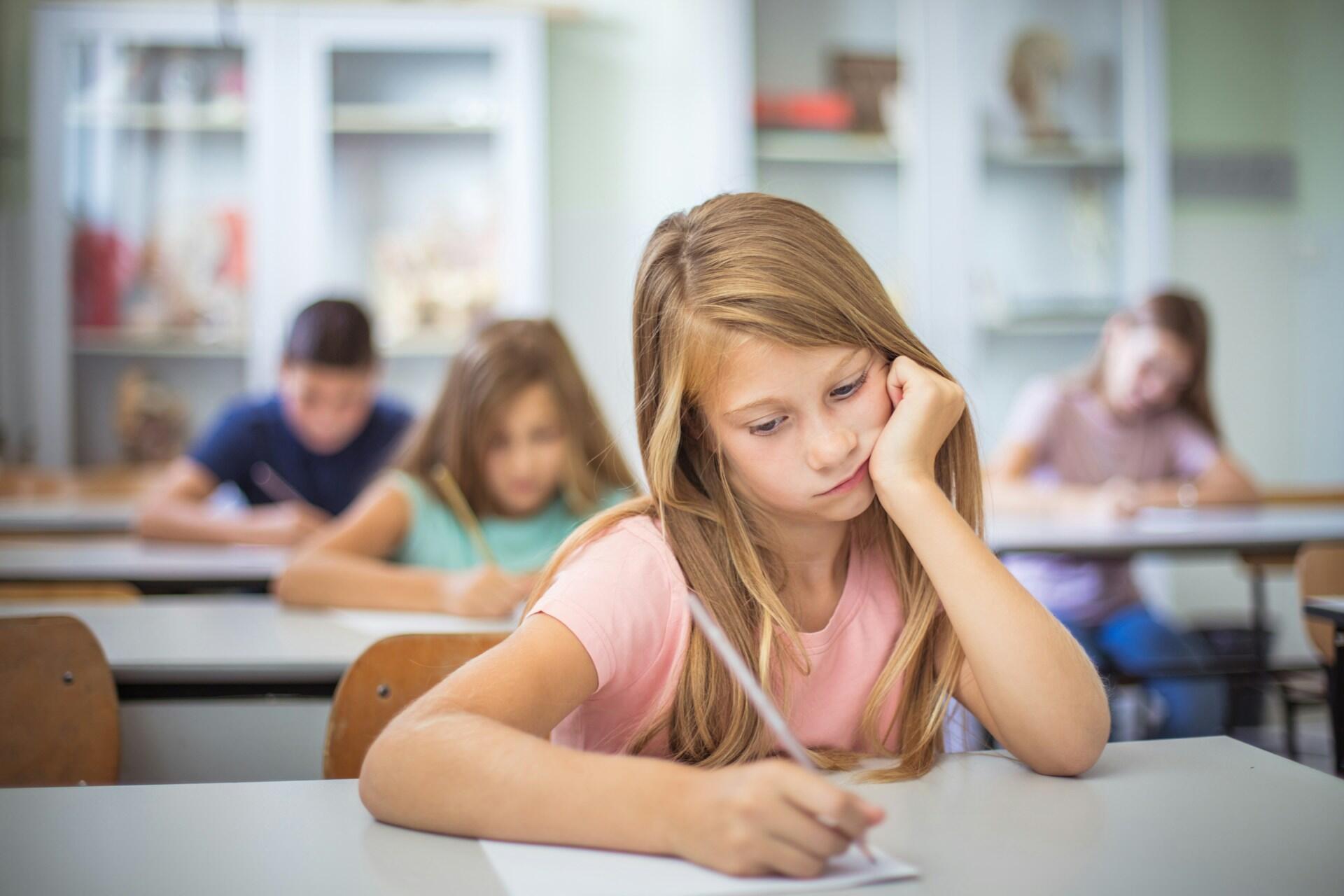 Une petite fille à l'école qui écrit sur une feuille et qui a l'air ennuyé